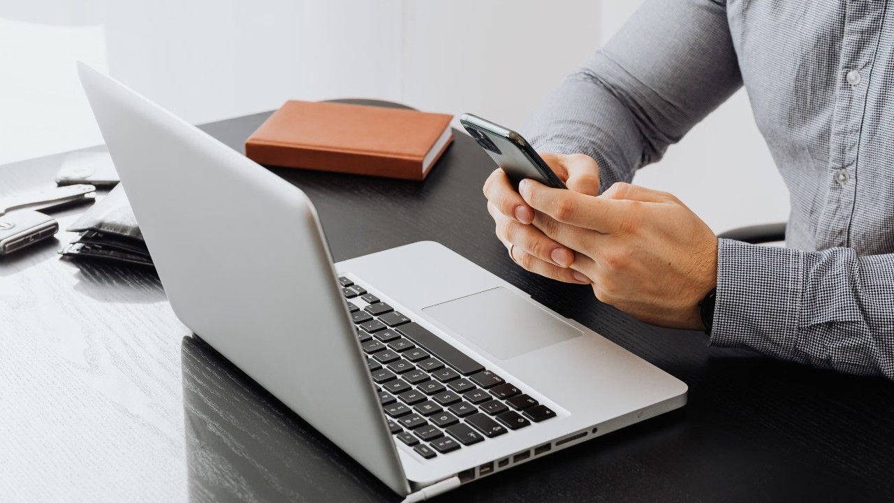 Mãos seguram celular ao lado de notebook sobre mesa escura